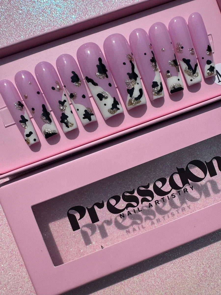 A set of long, pink press-on nails feature a white and black cow print design, accented with gold glitter. They have a glossy finish.