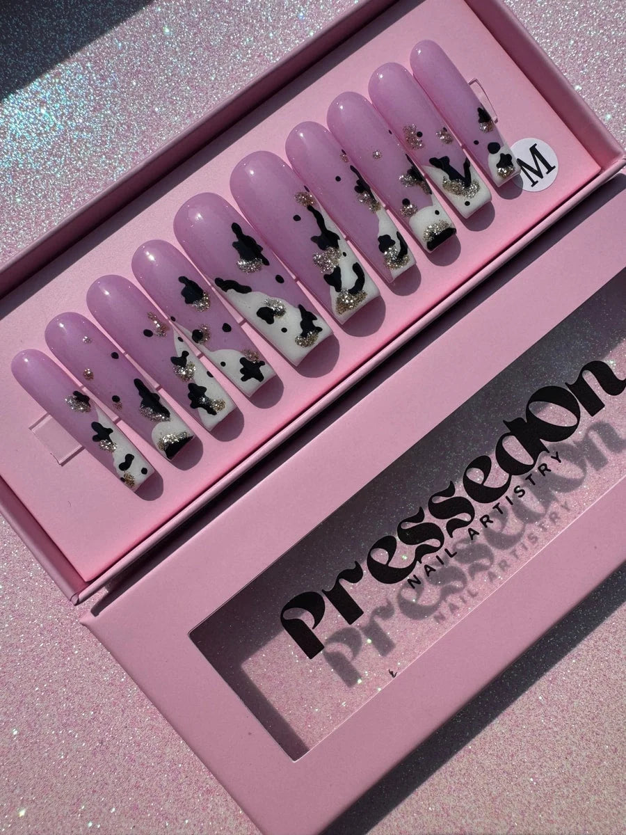 Long, coffin-shaped press-on nails with a light pink base and black cow print designs accented with silver glitter. 
