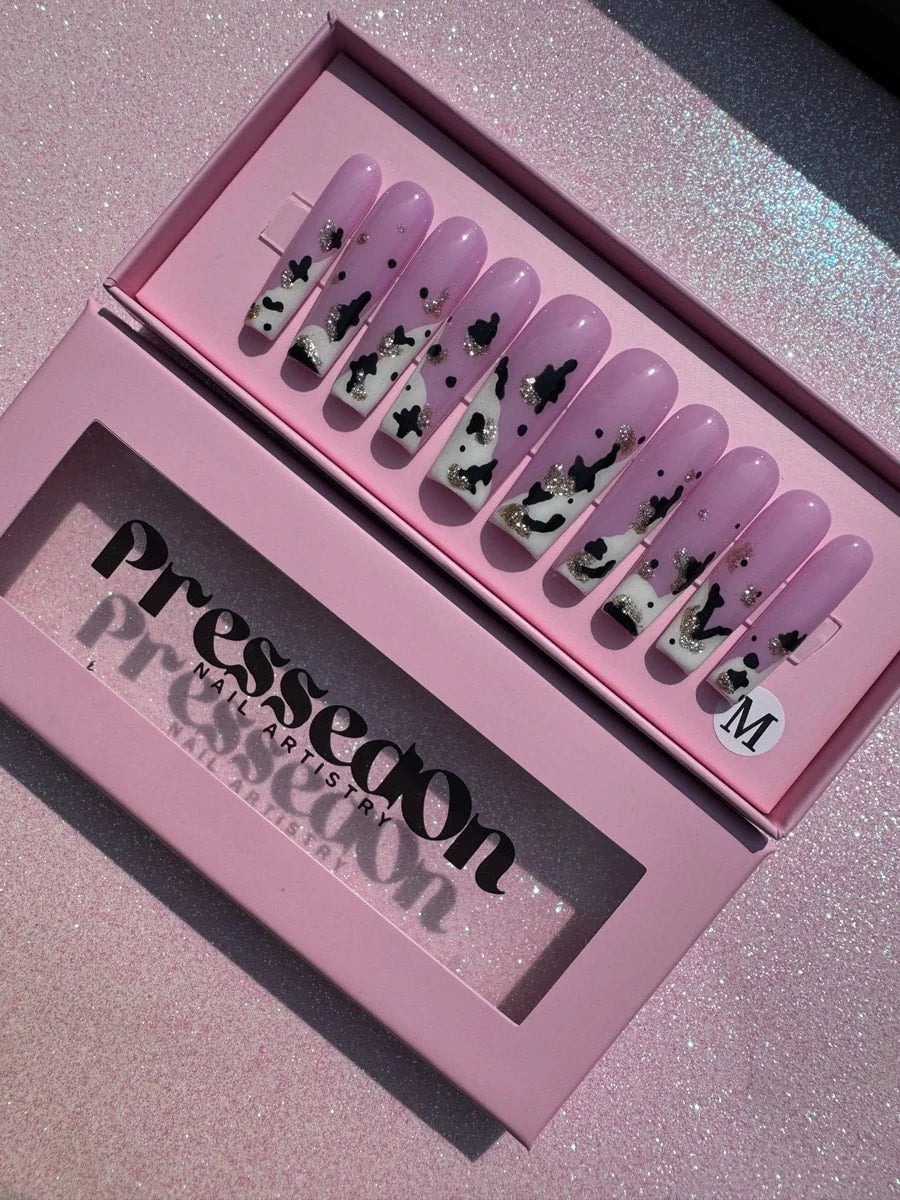 A set of ten long, coffin-shaped pink press-on nails features a cow print design with black and white spots and gold glitter.