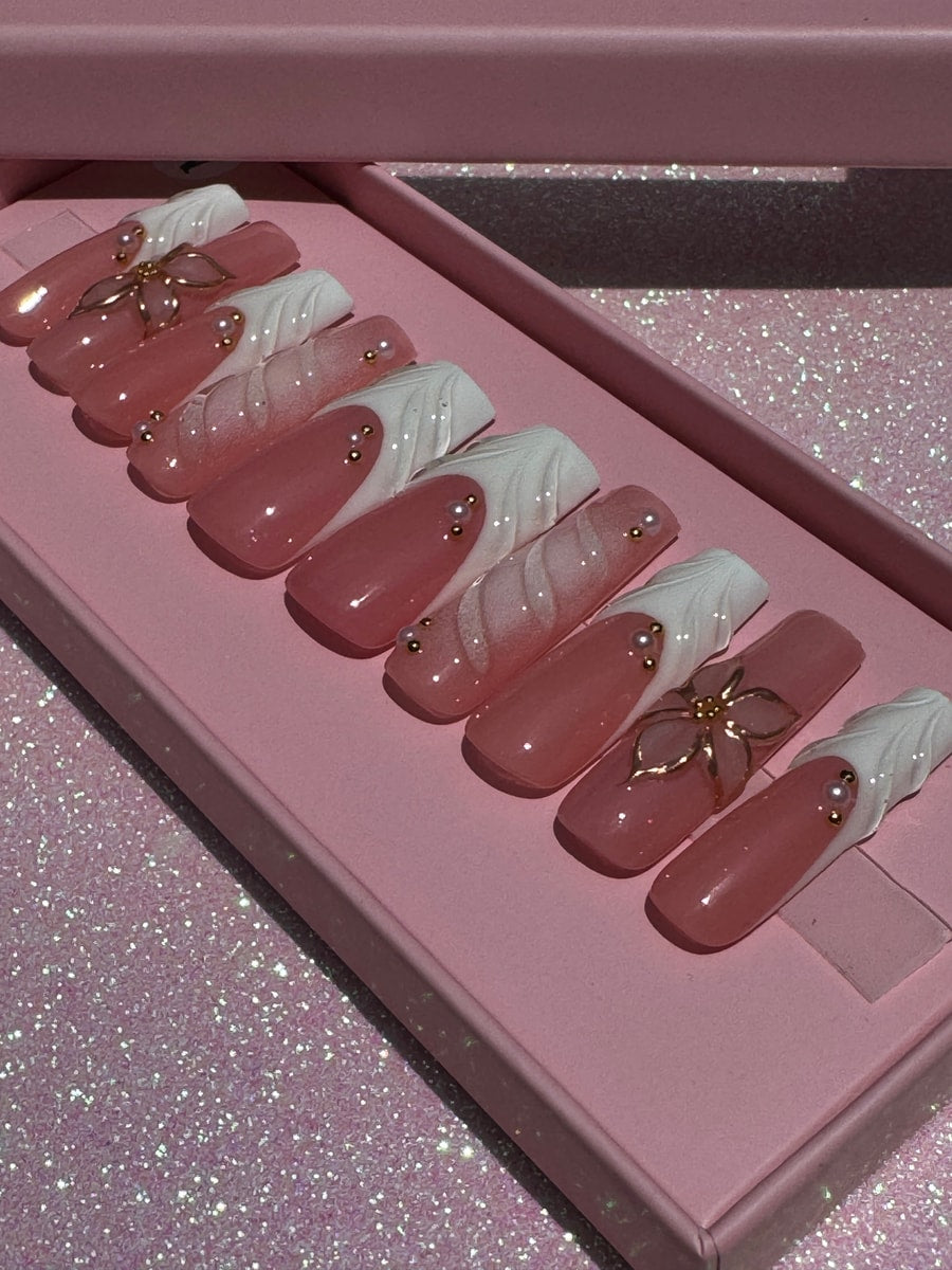 Pink and crystal clear press-on nails with white swirl accents and gold flower charms. Some nails feature small gold bead decorations.