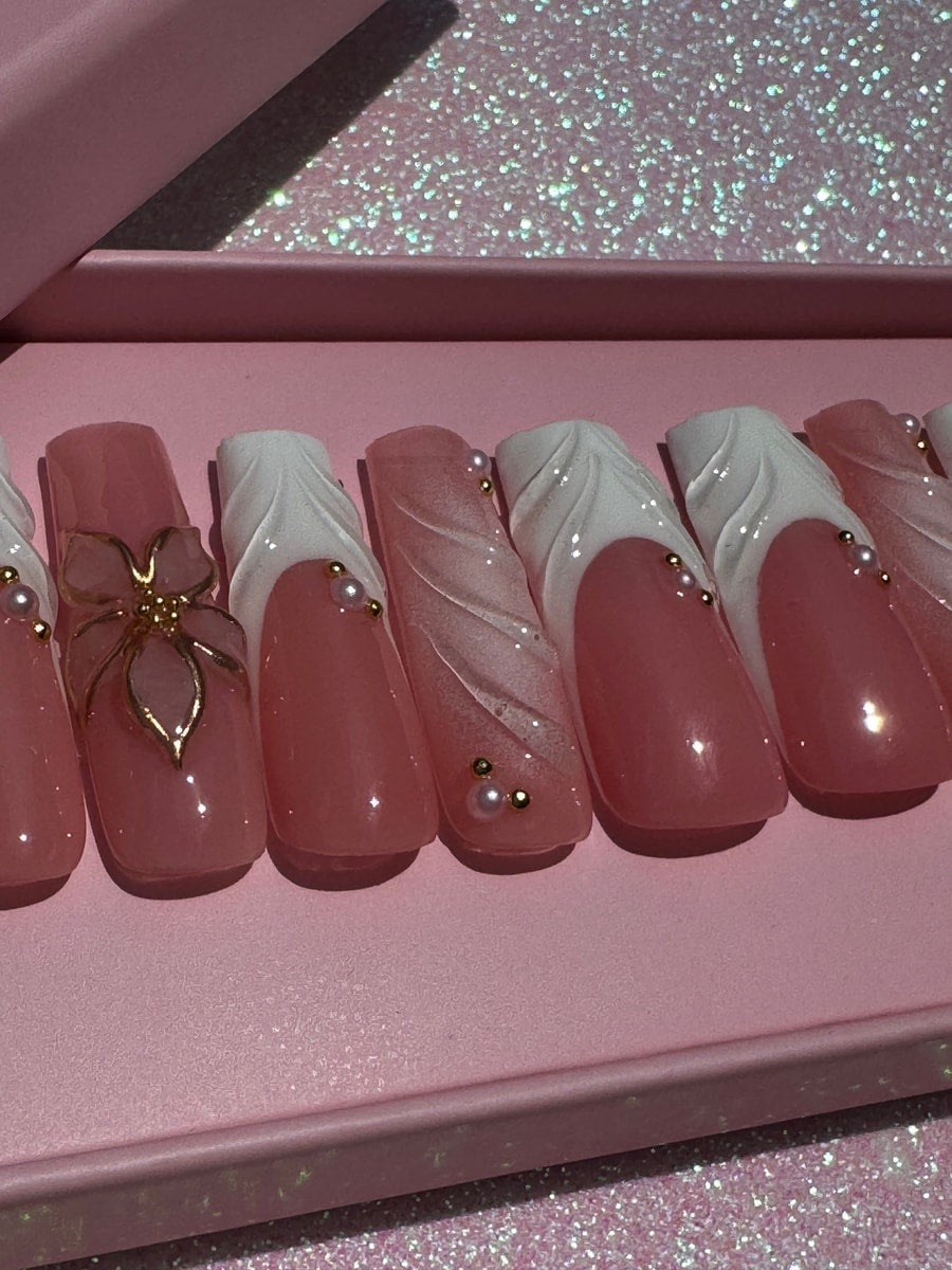 Pink crystal clear long press-on nails feature a white sculpted design and small gold bead and pearl accents. One nail has a gold flower charm.