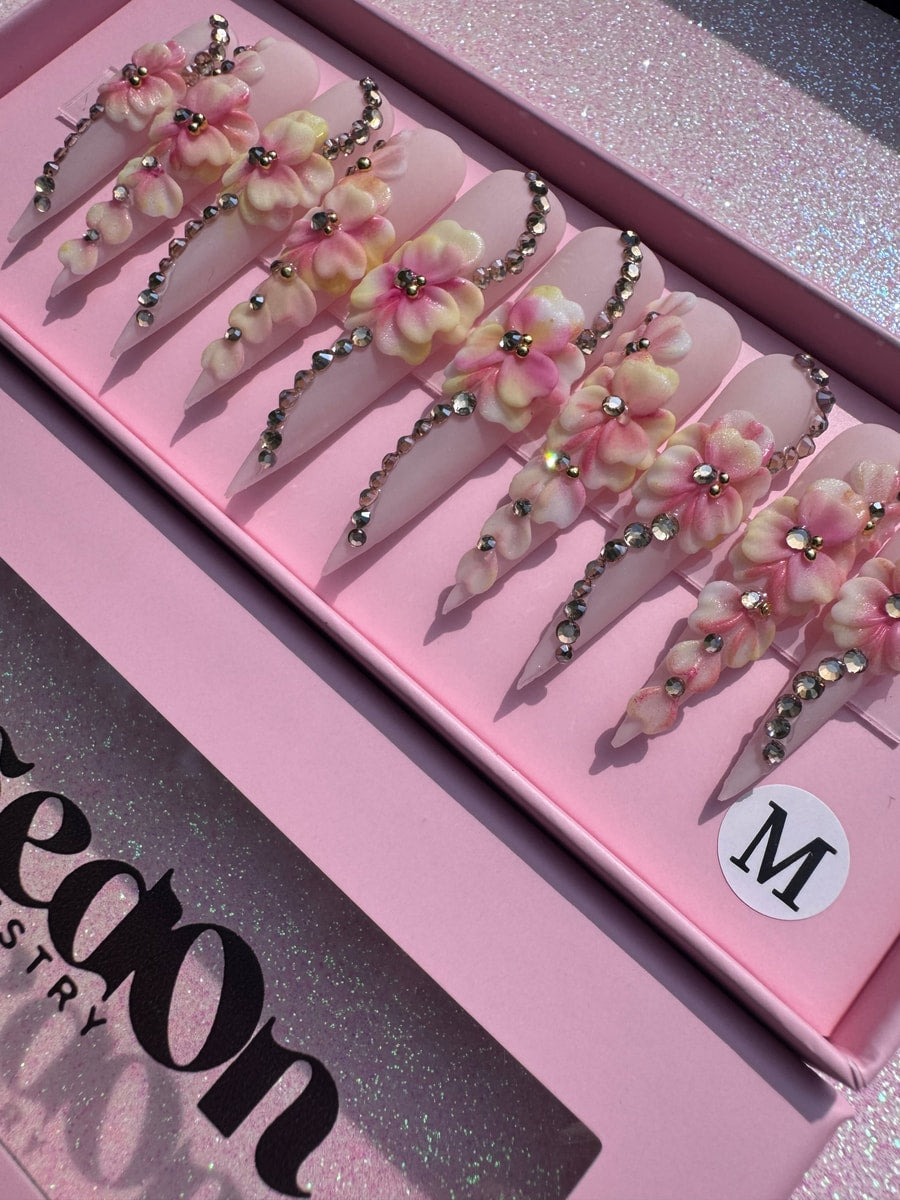 Light pink extra long stiletto press-on nails decorated with 3D flowers, gold accents, and a line of rhinestones running down the side.