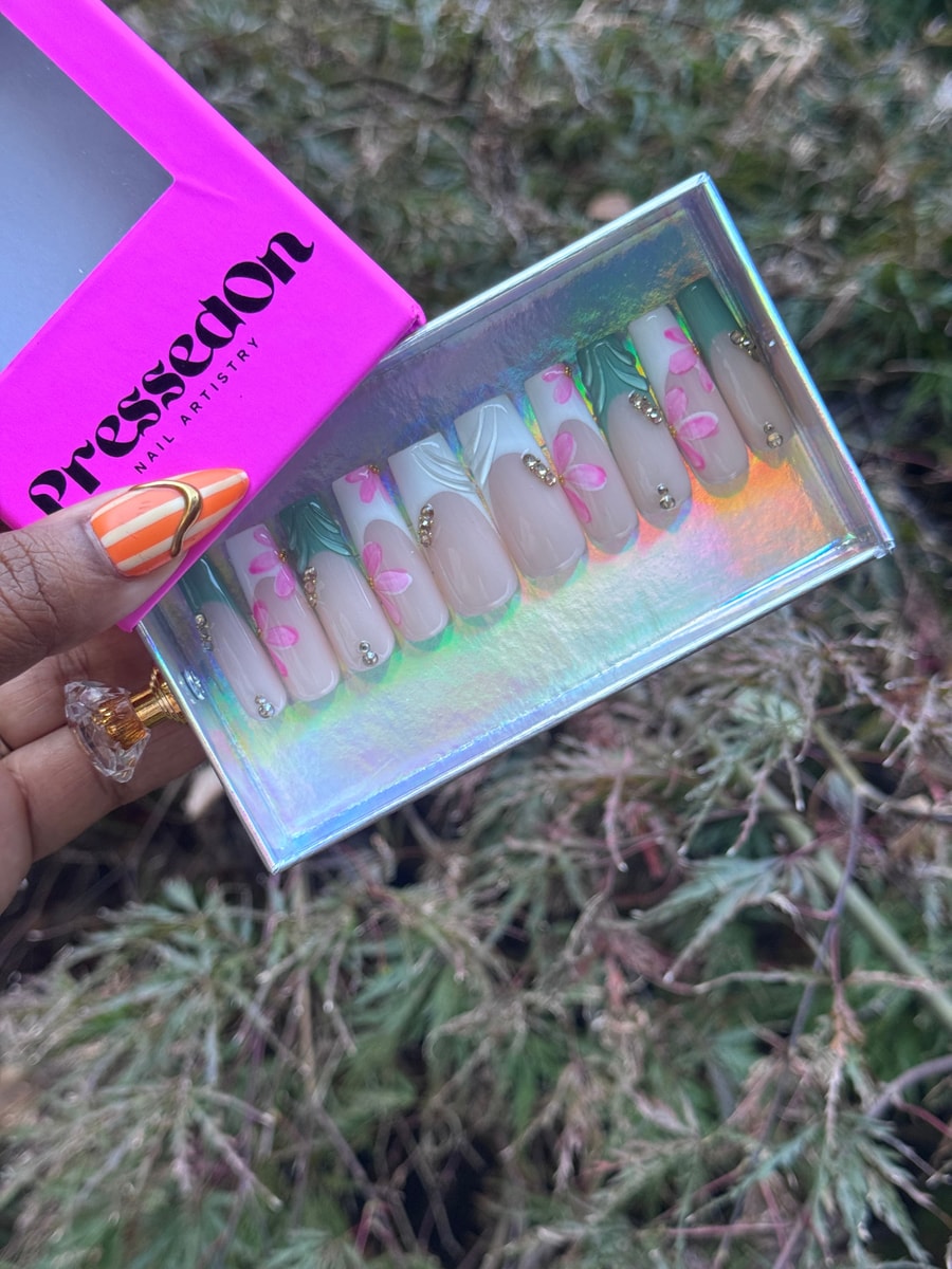 Medium-length press-on nails with a nude base, pink flower art, and white and green French tips. Each nail also has small rhinestone embellishments.