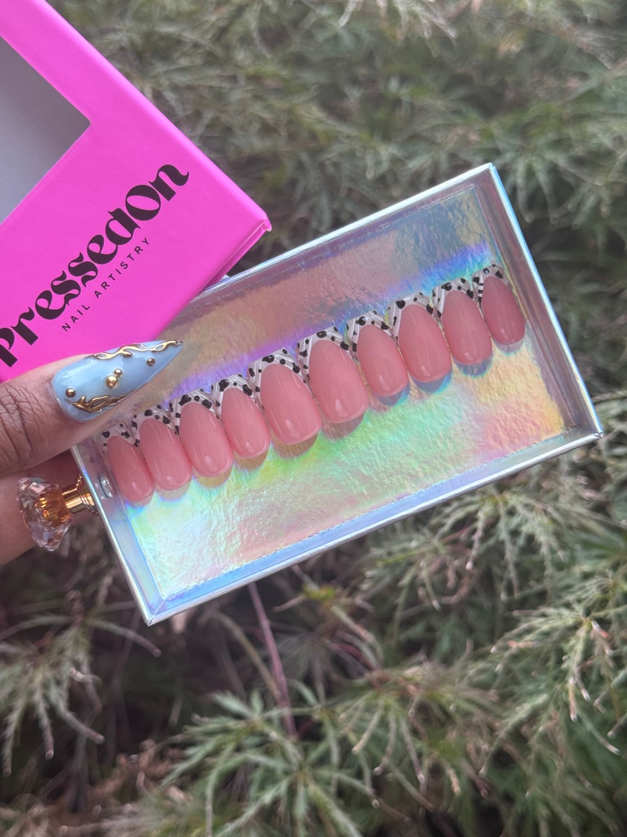 Short oval press-on nails with a light pink base and cow print French tips are displayed. The set is arranged in a holographic tray.