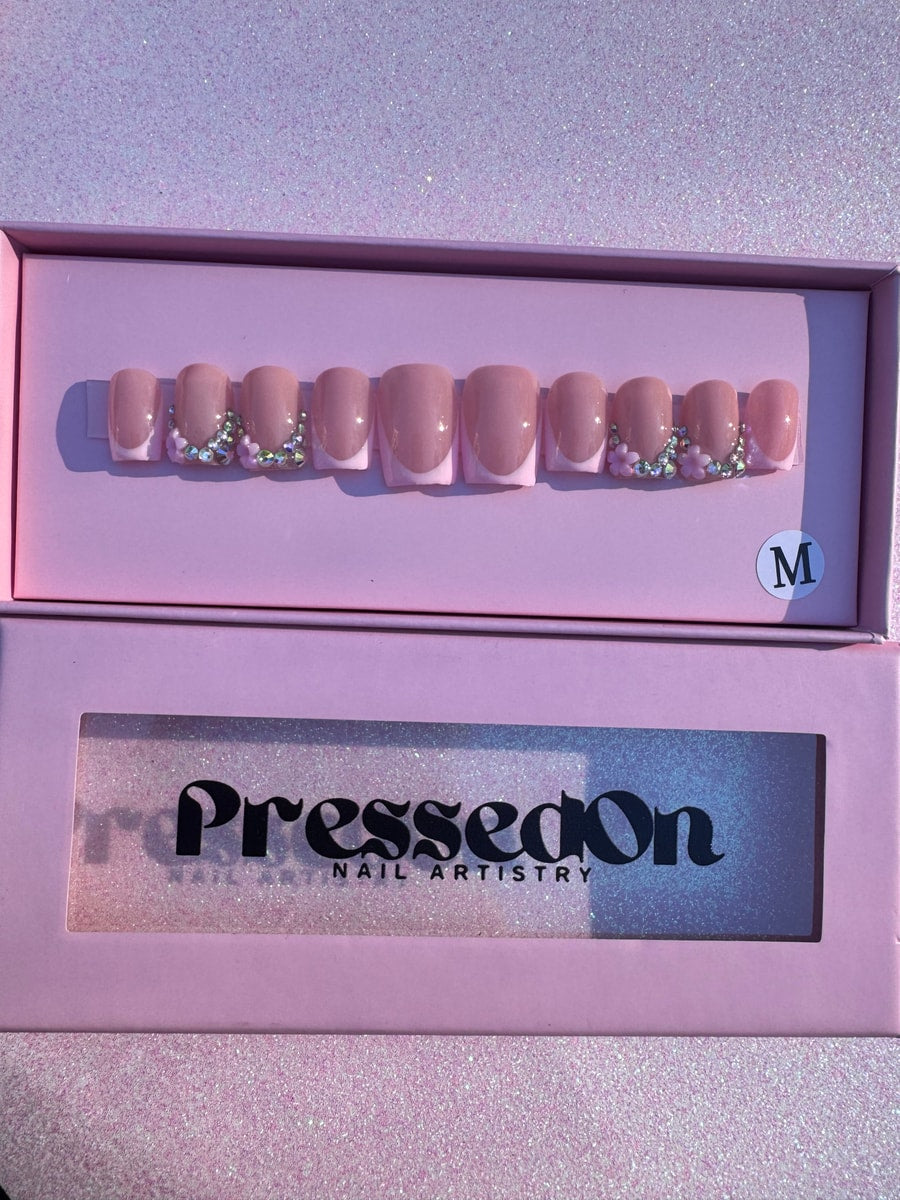 Super short pink press-on nails feature a white French tip design and sparkling bling accents with small flower charms.