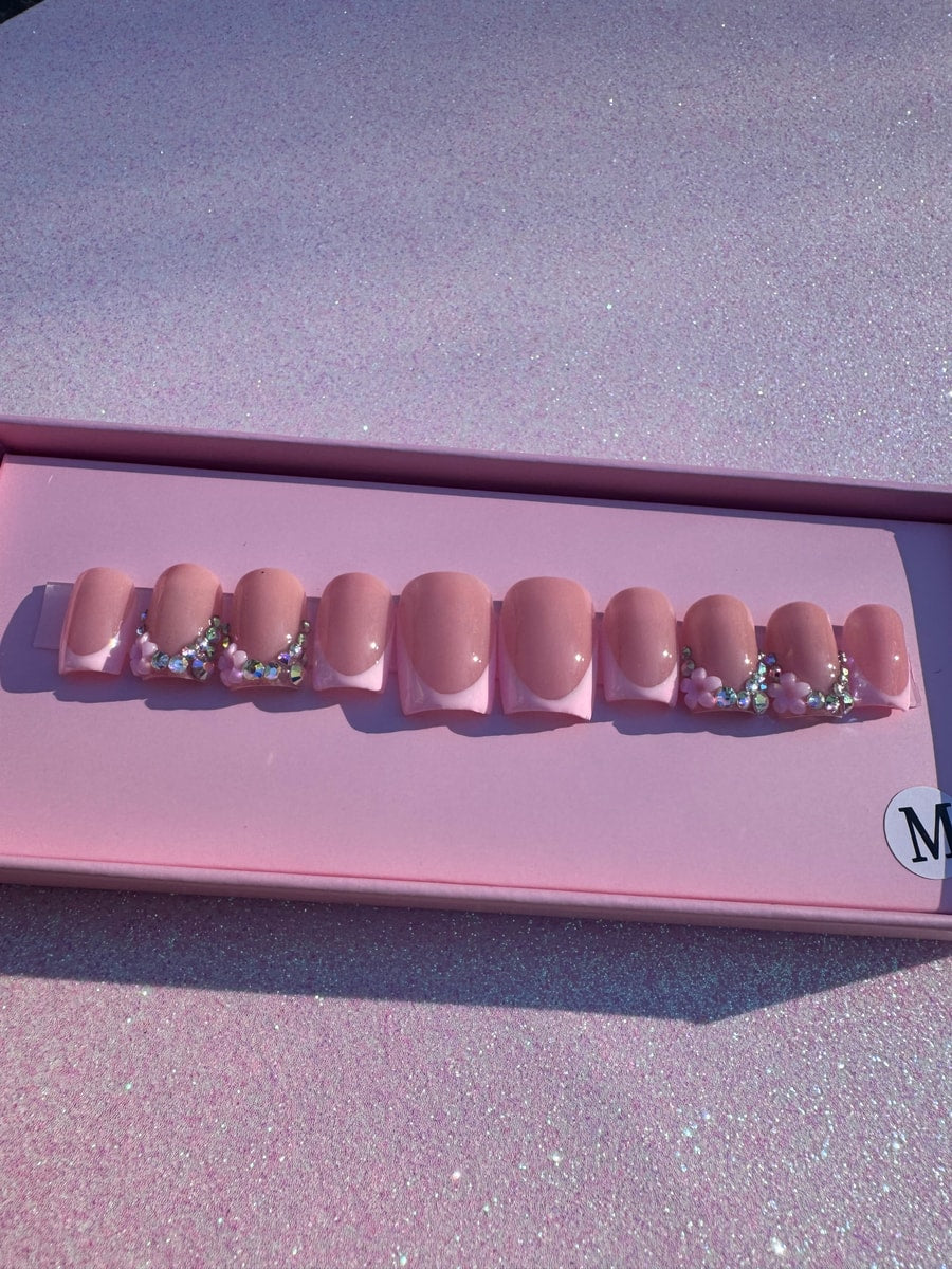 Short, square pink press-on nails feature a classic French tip design. Rhinestones and small flower charms adorn some of the nails.