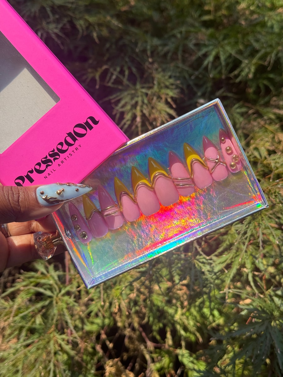 The image displays short stiletto press-on nails with a pink base and gold tips. They have gold wire accents, and one nail features three gold embellishments.