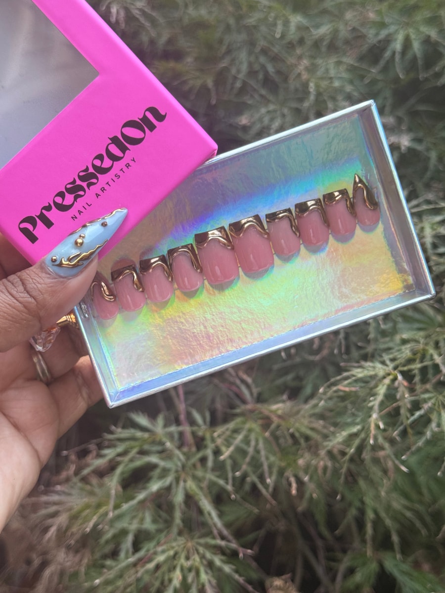 Short, square pink press-on nails with gold accents and a drip design. One nail features a blue base with gold embellishments.