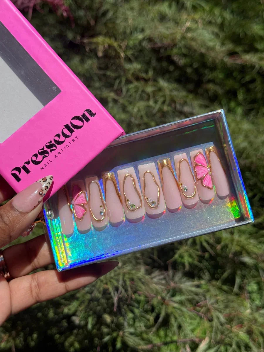 Medium pink press-on nails with gold accents and delicate half hibiscus flower charms. Several nails also have small rhinestone details.