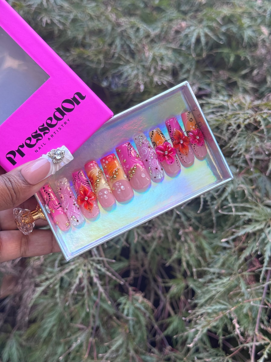 Pink island bloom press-on nails with orange floral accents and gold rhinestones. The nails have a medium length and various unique embellishments.