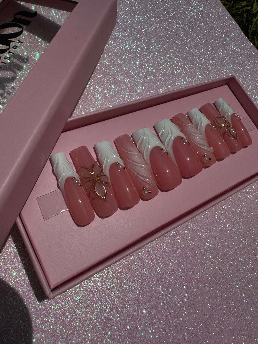 Long pink press-on nails with a white swirl design on the tips. Several nails are decorated with gold flower charms and small pearl accents.