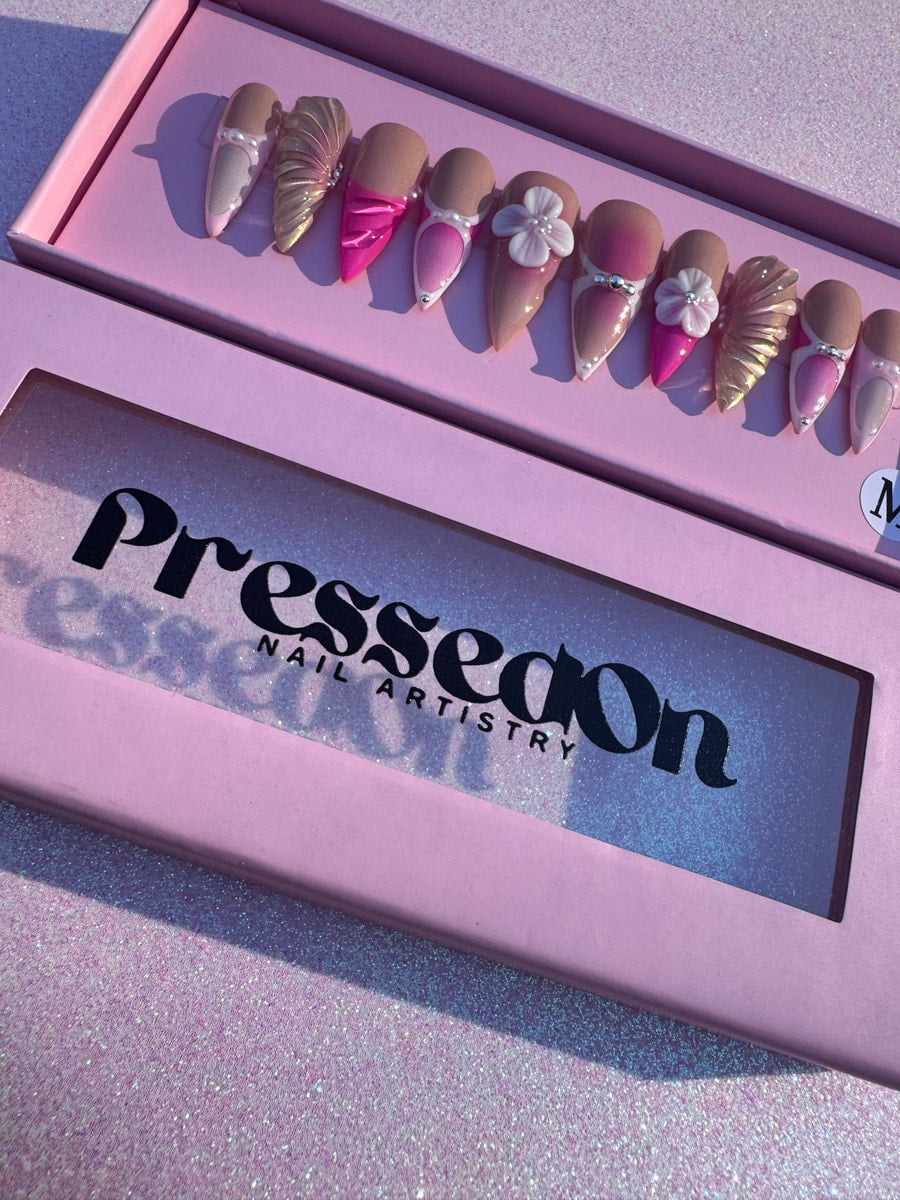 A set of medium-length press-on nails with a mermaid-inspired design, featuring pink and nude shades, 3D shell accents, white floral designs, and pearl details.