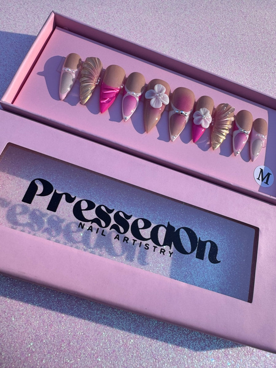 A set of pink mermaid press-on nails in medium length featuring pearl accents, a 3D flower, and gold shell details.