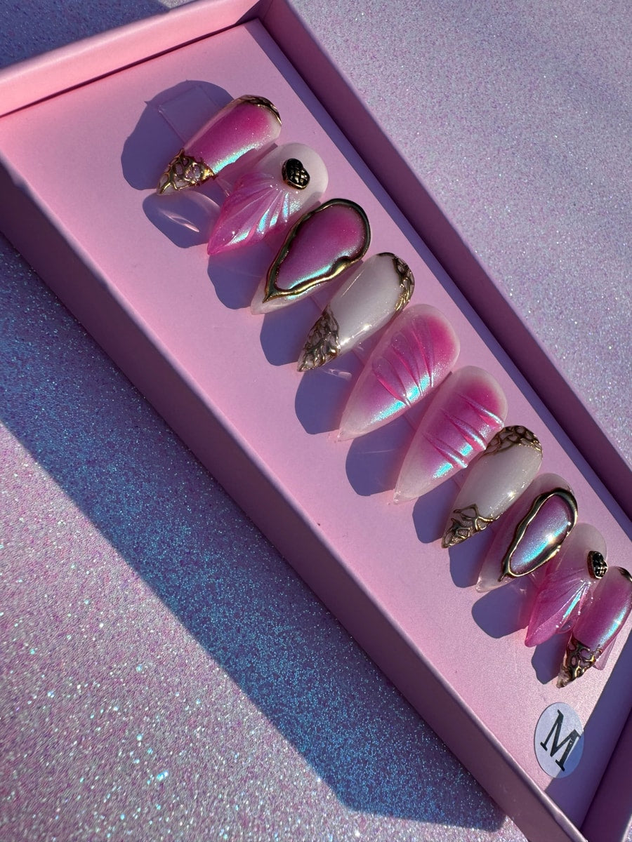 A set of medium-length pink paradise press-on nails features pink, white, and gold color schemes. Some have solid color, while others feature unique designs and iridescent effects.