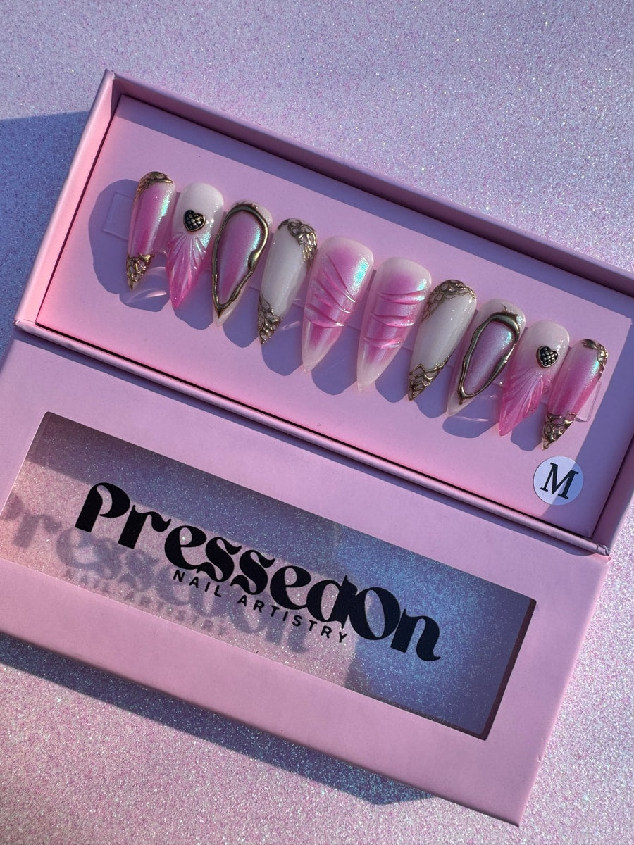 Medium length pink and white stiletto press-on nails featuring pearlescent finish, gold accents, and heart charms. Some nails have pink leaf designs and gold outlining.