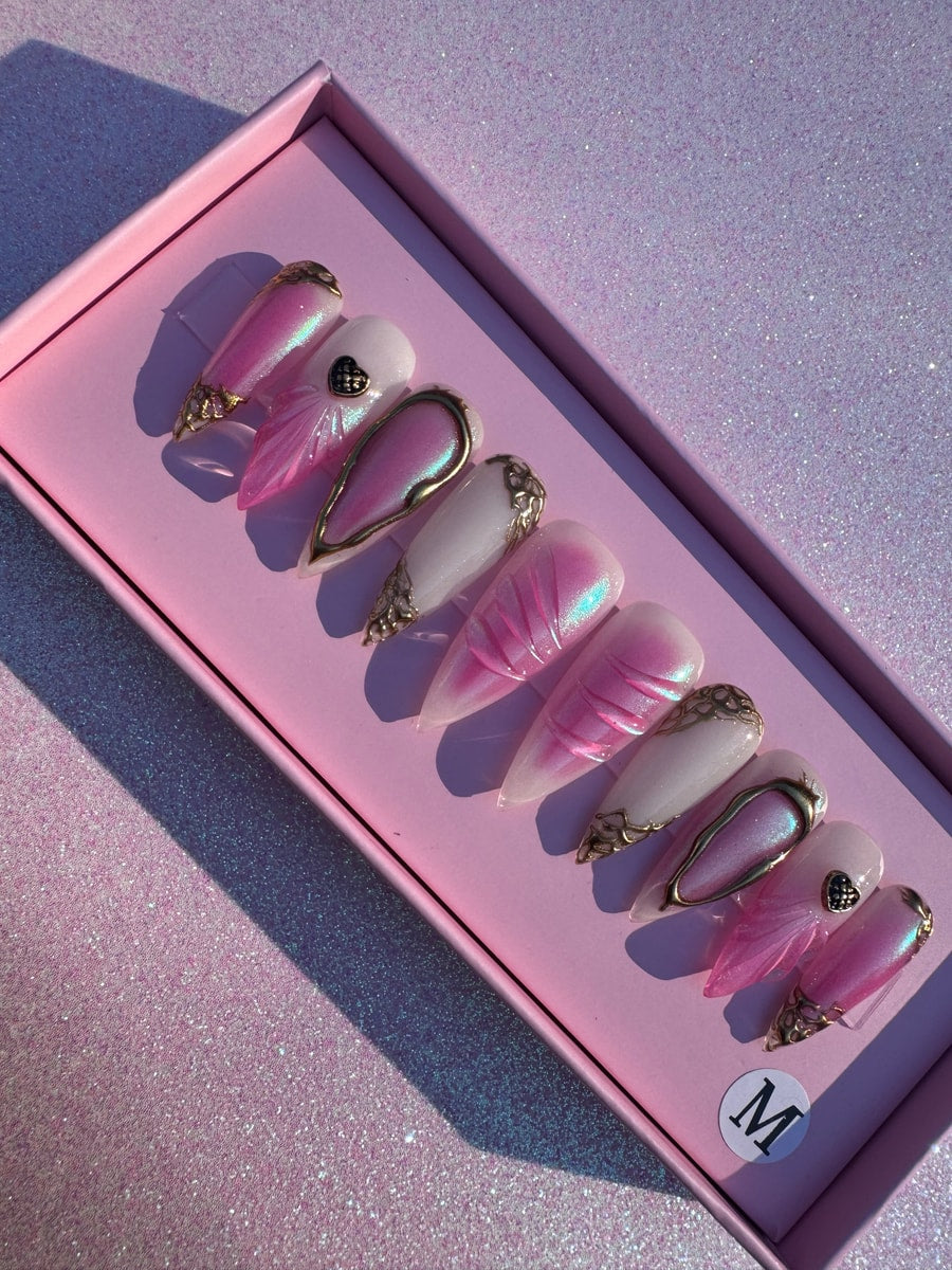 A set of medium-length stiletto press-on nails. Designs include iridescent pink, solid white, gold filigree accents, and a small black heart charm.