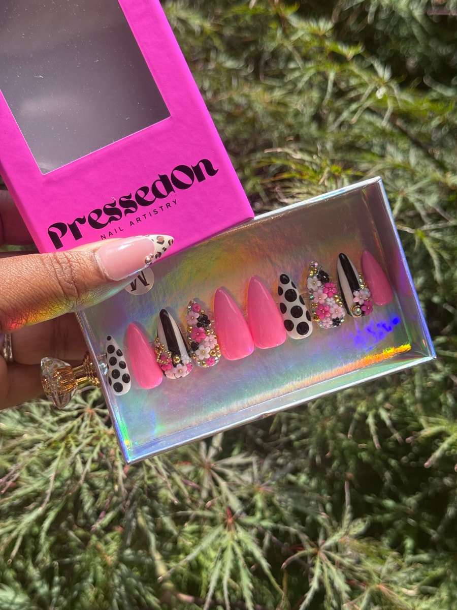 Almond-shaped press-on nails with pink, black, and white polka dot patterns, accented with 3D flowers and gold gems; also includes solid pink nails.