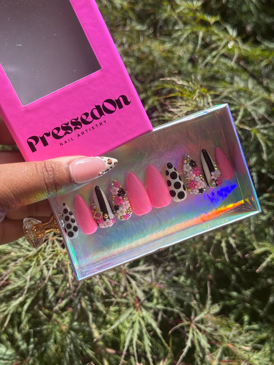 Set of short, stiletto shaped pink press-on nails. Several nails have polka dot patterns and 3D floral charm accents in white, black, and gold.