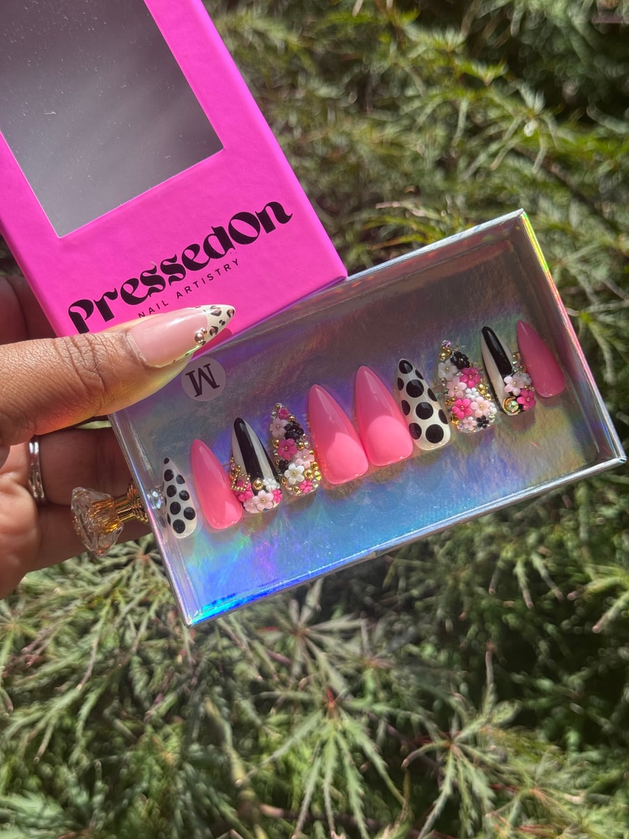 A set of short stiletto press-on nails including pink, white with black polka dots, and black with white stripe designs accented with 3D flowers and gold studs.