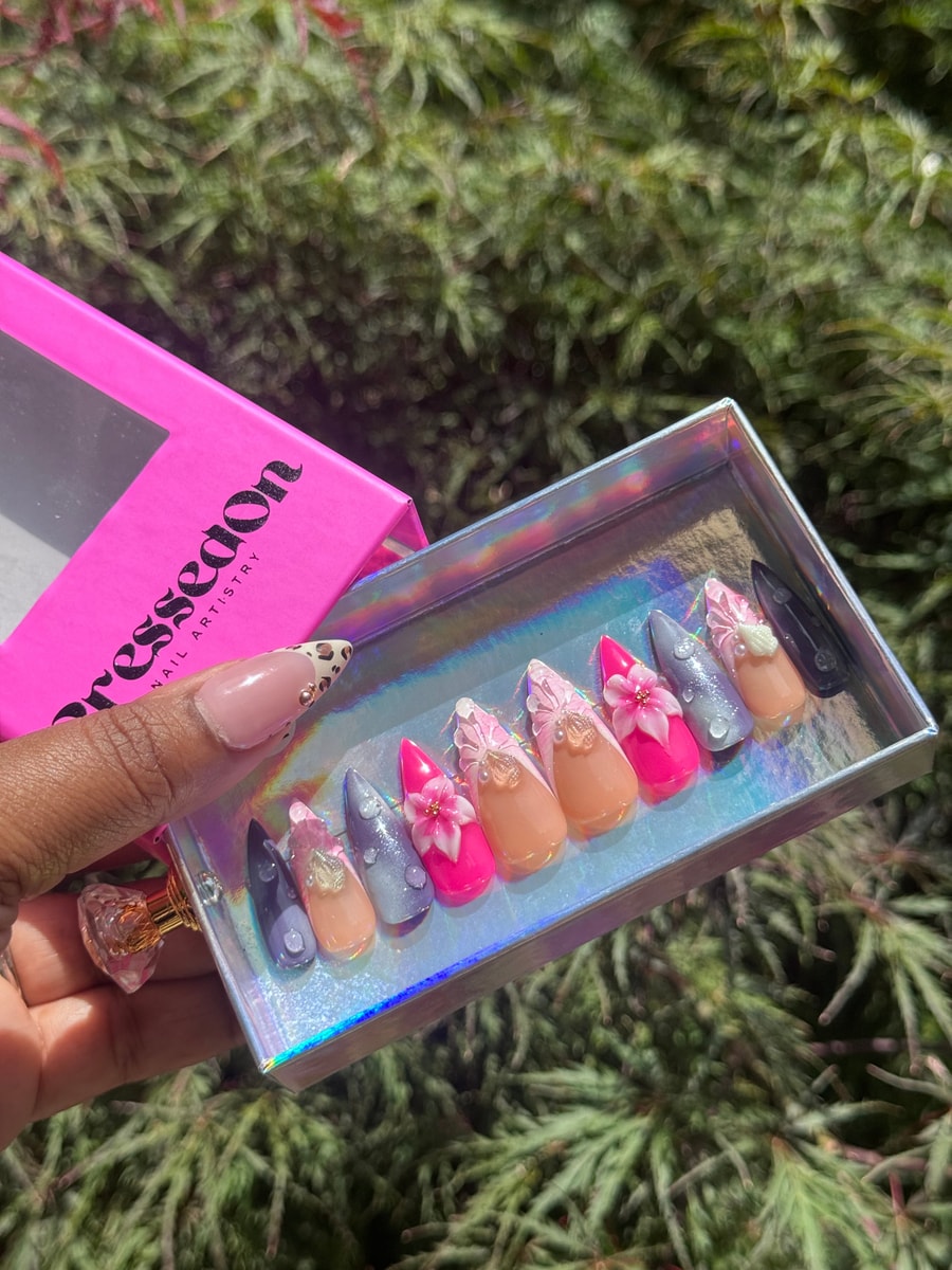 Medium pink press-on nails featuring 3D flower decorations, iridescent details, and a mix of pink, gray, nude, and black designs.