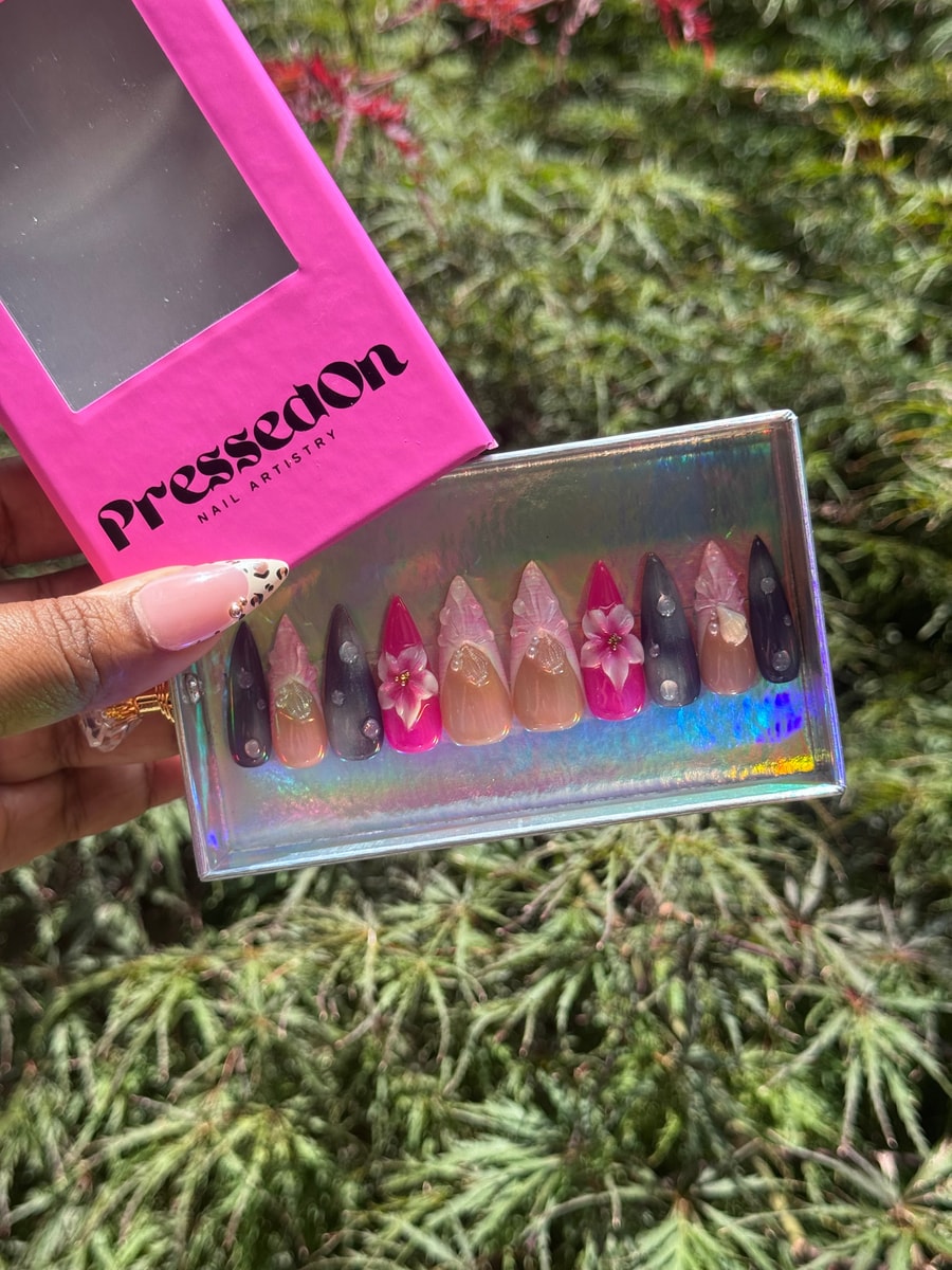Medium-length press-on nails in a pink pop color scheme with black and nude accents. Each nail is decorated with 3D flower charms and iridescent gems.
