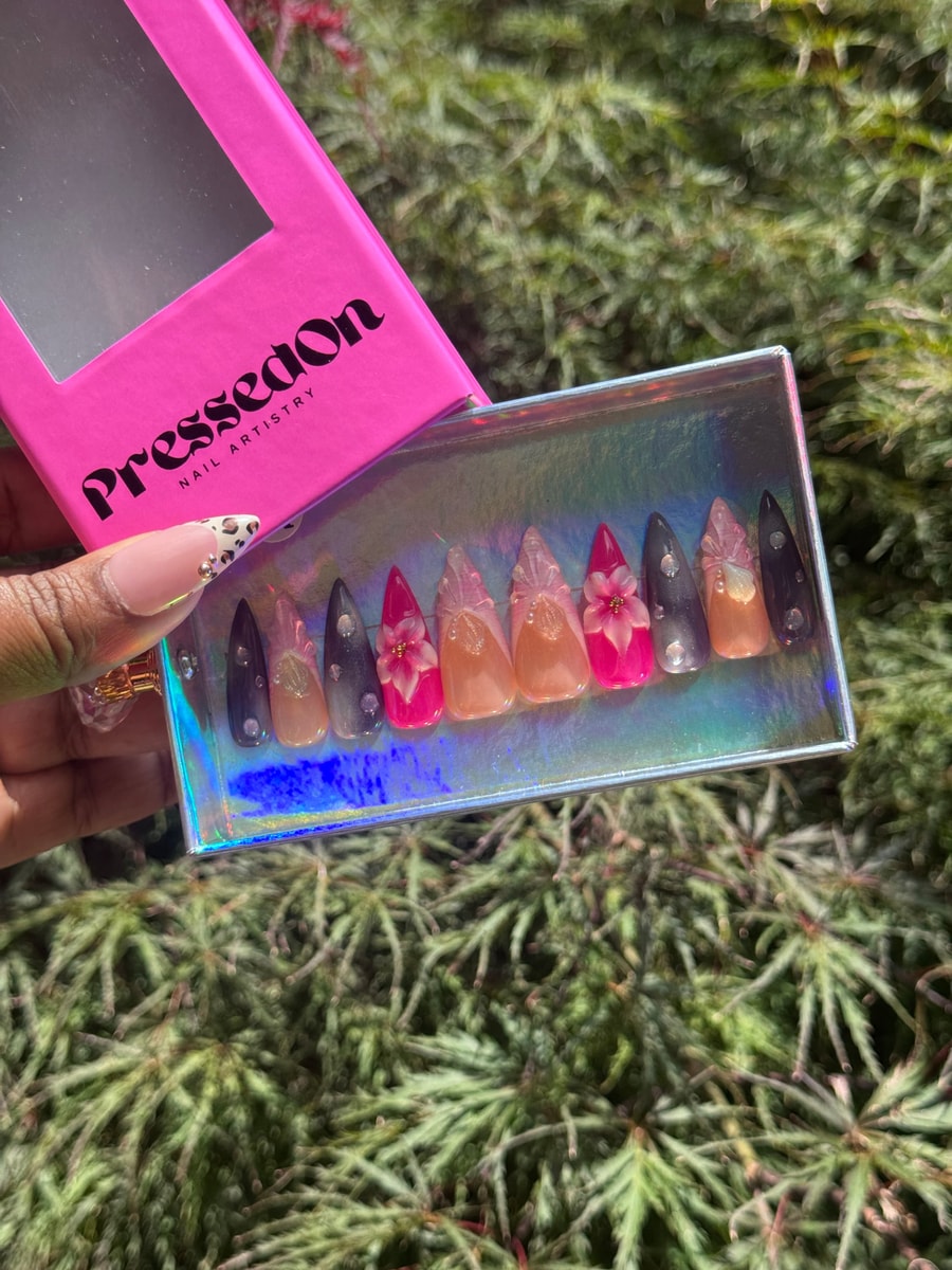 Medium length stiletto press-on nails with flower embellishments and rhinestones in pink, purple, and nude shades.