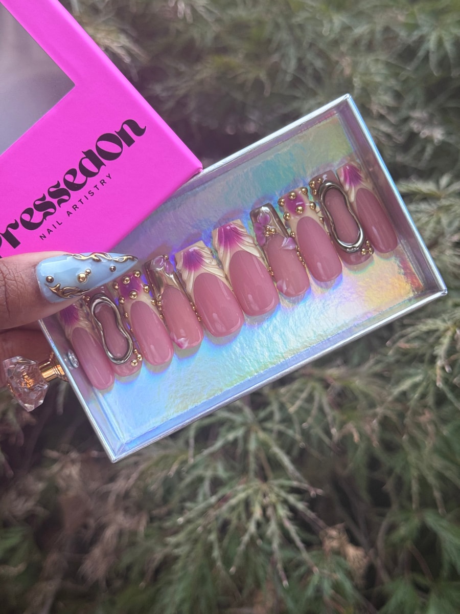 Short pink press-on nails with gold details, purple floral accents on white tips, and one blue accent nail with gold embellishments. The set has a feminine, artistic design.