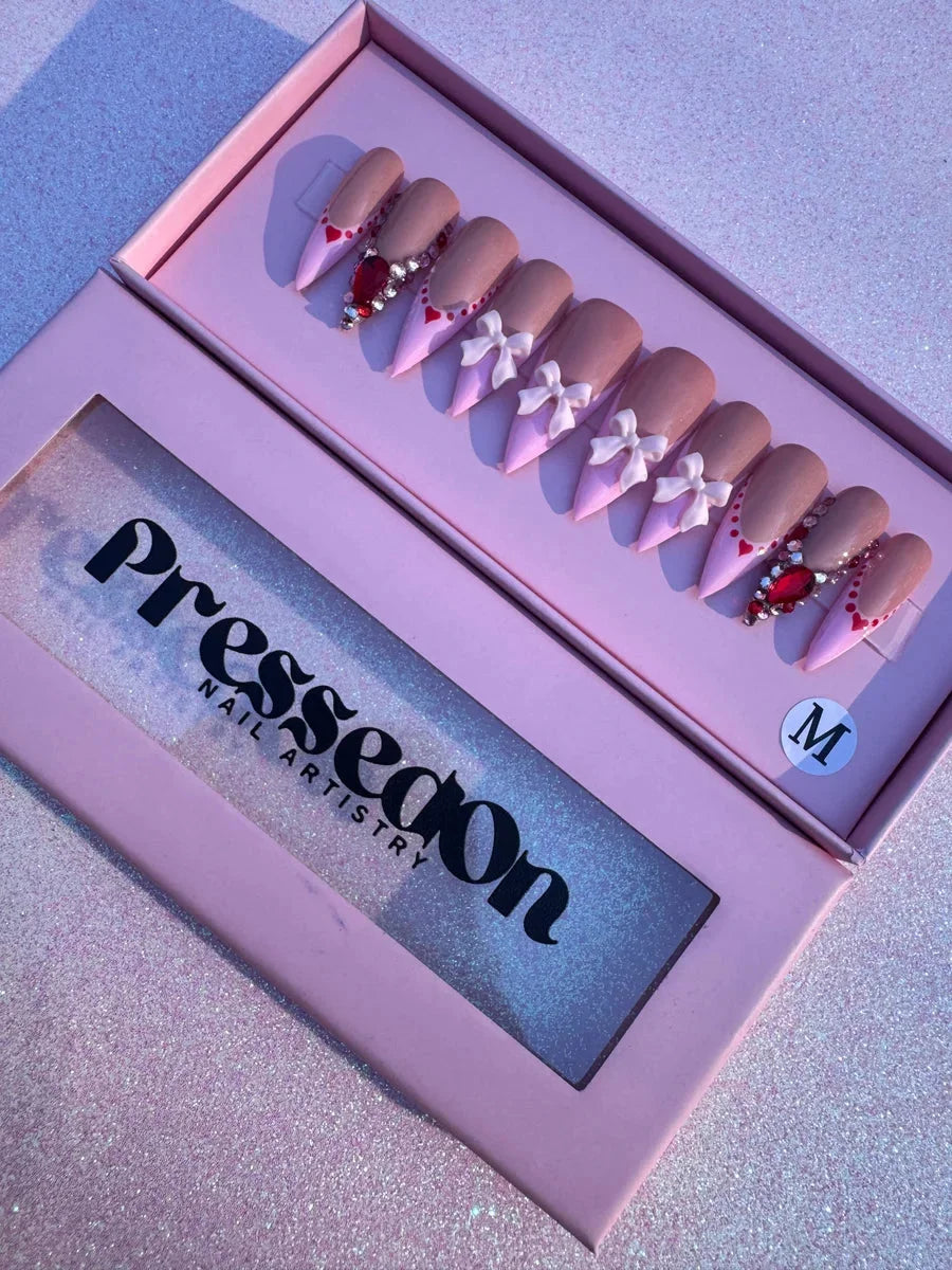 Valentine's Day themed stiletto press-on nails with nude and pink color blocking, embellished with bows and heart-shaped gemstones.