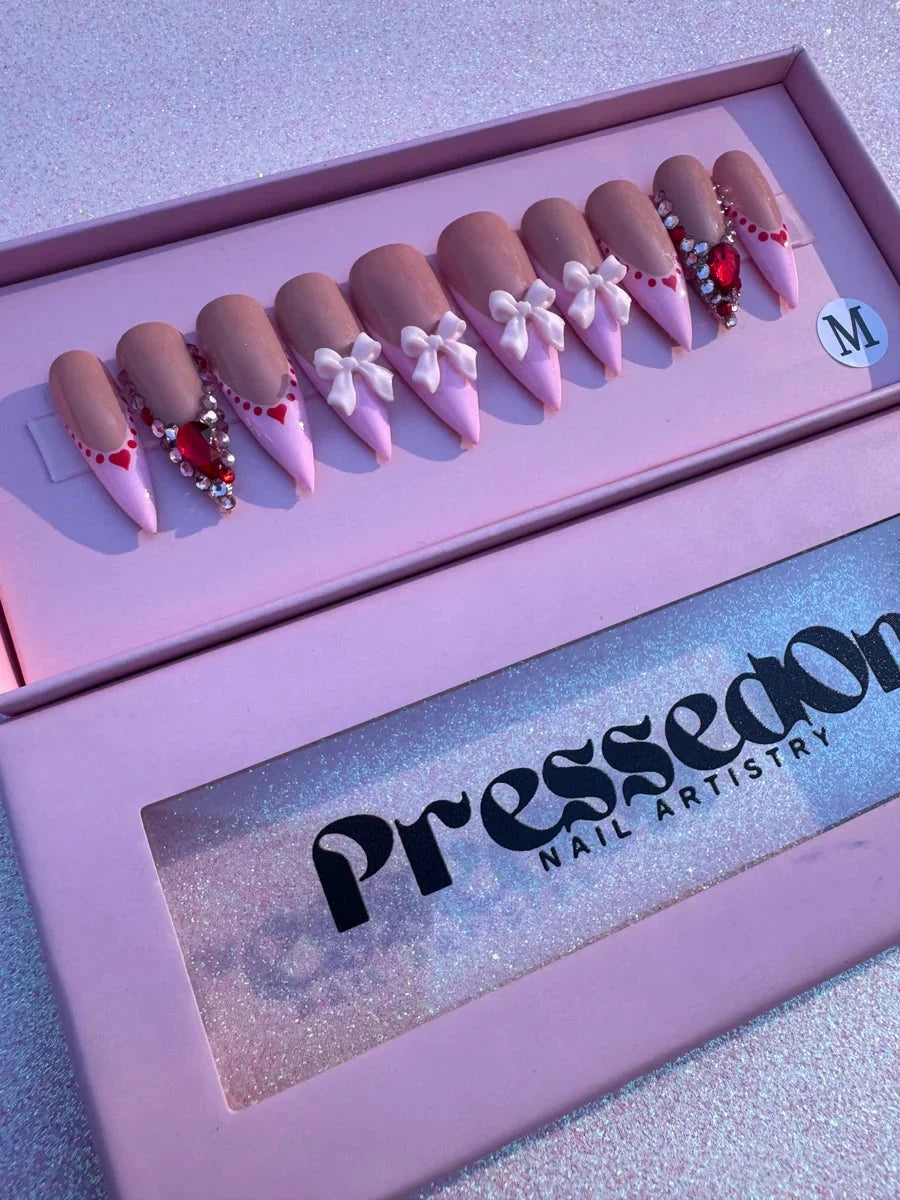 Pink stiletto press-on nails with nude bases, pink tips, adorned with white bows, red heart details, and sparkling gemstones.