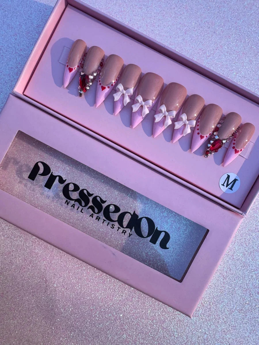 Medium-length nude and pink press-on nails featuring heart accents, gemstone embellishments, and bow decorations.