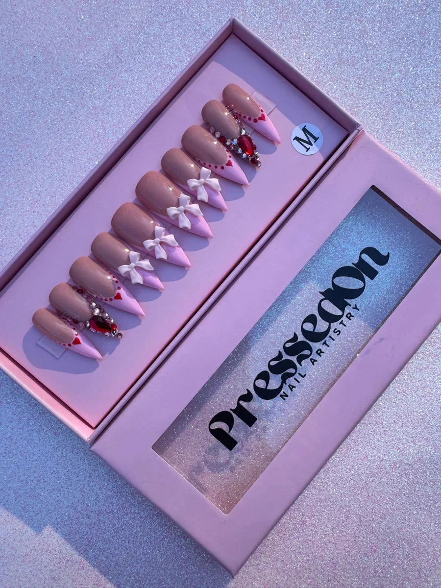 Pink and nude medium press-on nails featuring pink tips, white bows, and red gemstone accents for a Valentine's Day design.