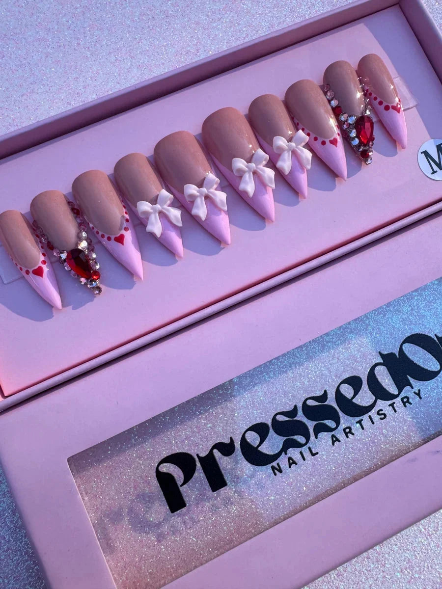 Pink stiletto press-on nails with nude accents, decorated with white bows, red gemstones and hearts for a Valentine's Day design.