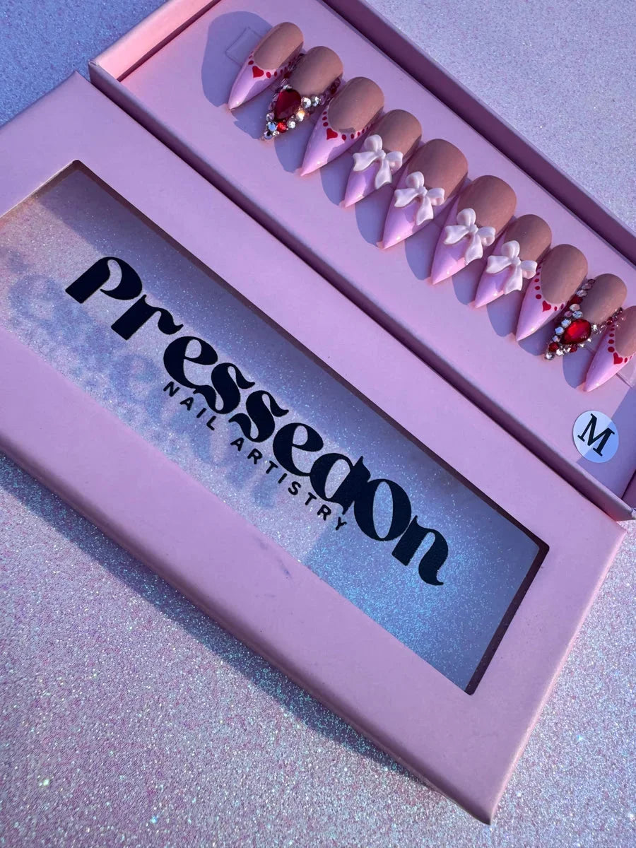 Valentine's Day press-on nails with a nude and pink design, bows, and gemstones.
