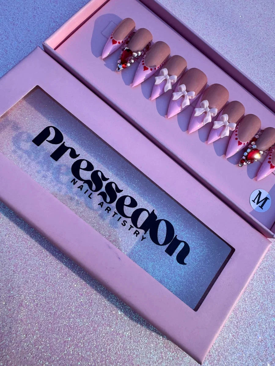 Medium-length press-on nails for Valentine's Day, featuring a pink base, tan tips, white bows, small hearts, rhinestones, and a large red gemstone accent.