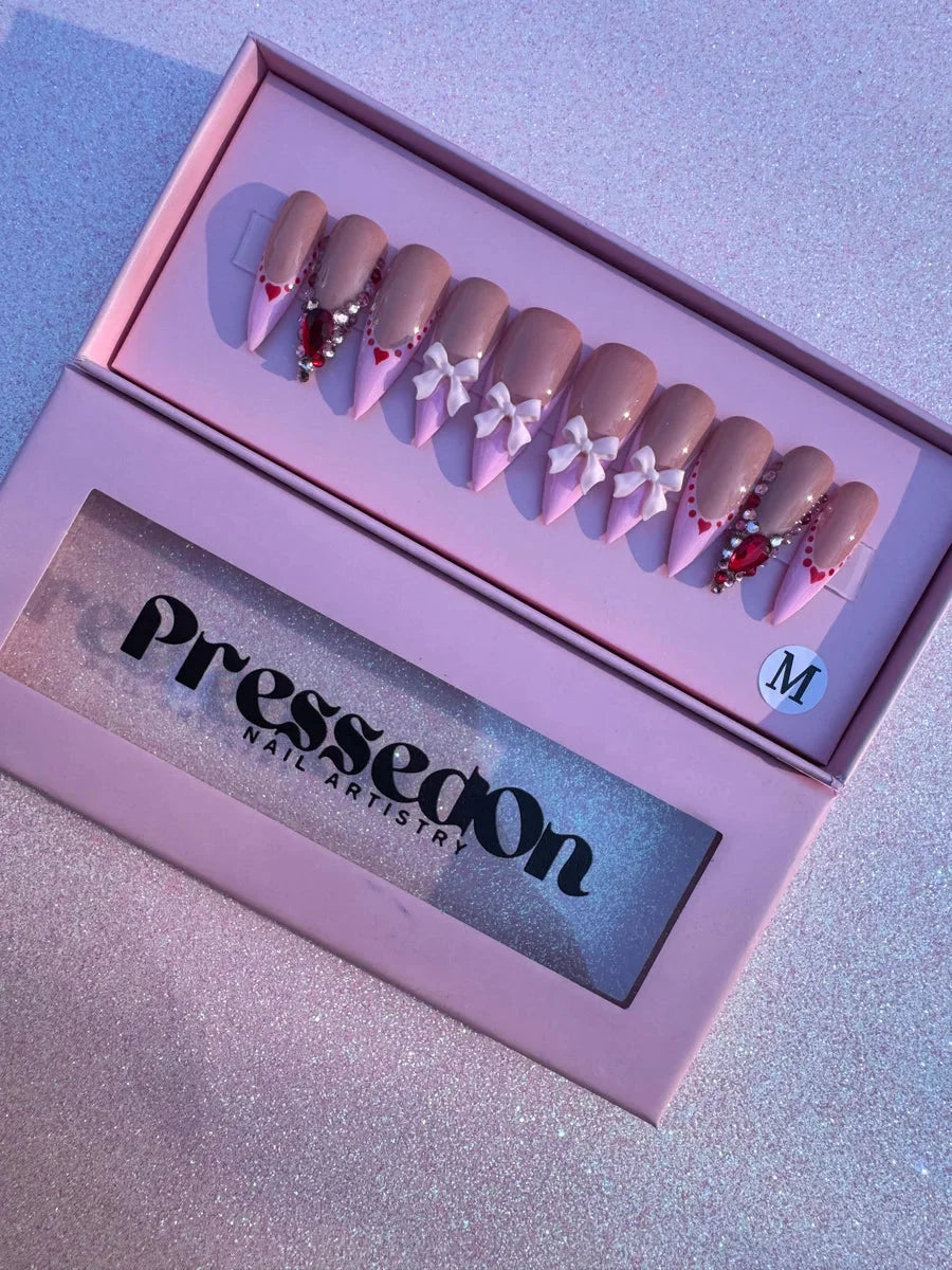 Medium pink press-on nails for Valentine's Day. They have a pink base, heart accents, 3D bows, and gemstone embellishments.