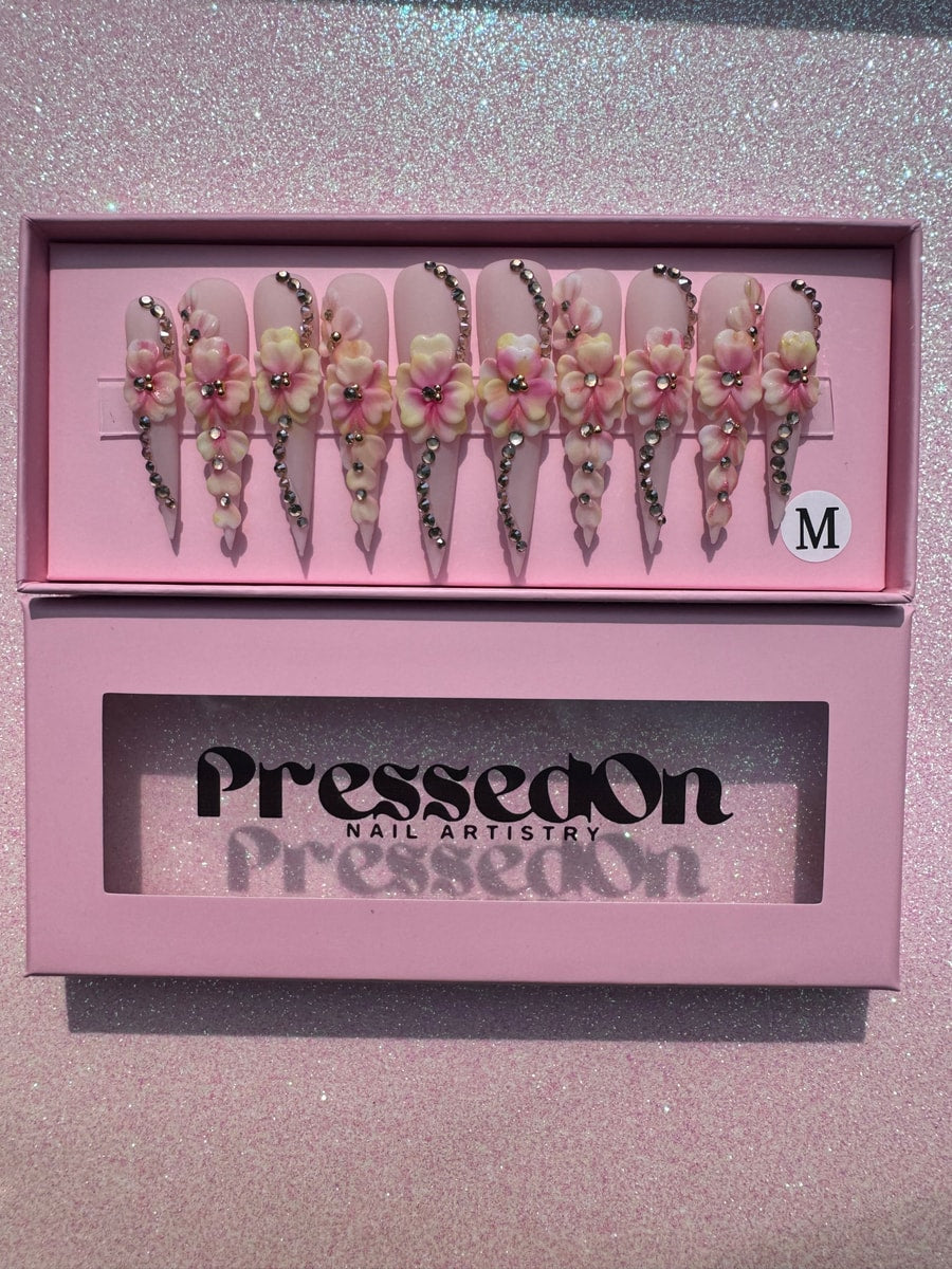 These are pink stiletto press-on nails with a 3D floral design and rhinestone accents. The nails are extra long and have a glossy finish.