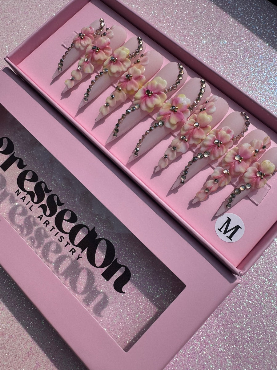 Extra long stiletto press-on nails in a pink and white color scheme with 3D flower embellishments and rhinestone details.