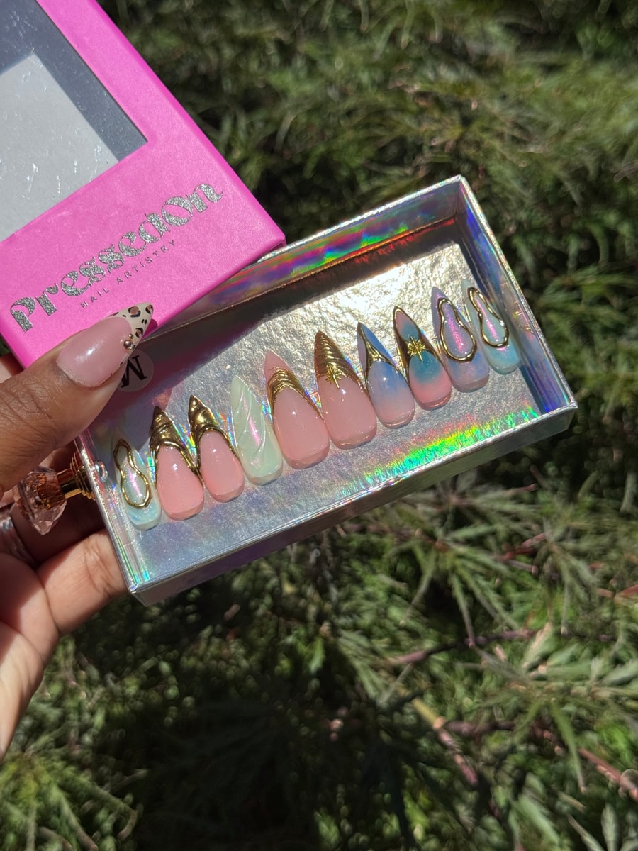 This set of medium stiletto press-on nails features pink, blue, and iridescent nails with gold details. Several nails are embellished with gold elements and charms.