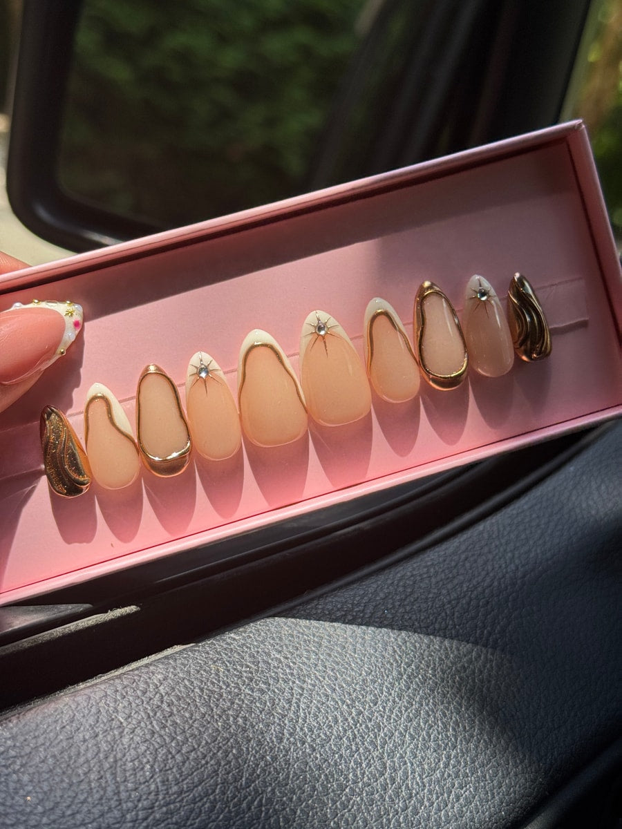 Set of princess super short almond-shaped press-on nails with nude, white, and gold abstract designs. Some nails feature rhinestones and gold wavy patterns.