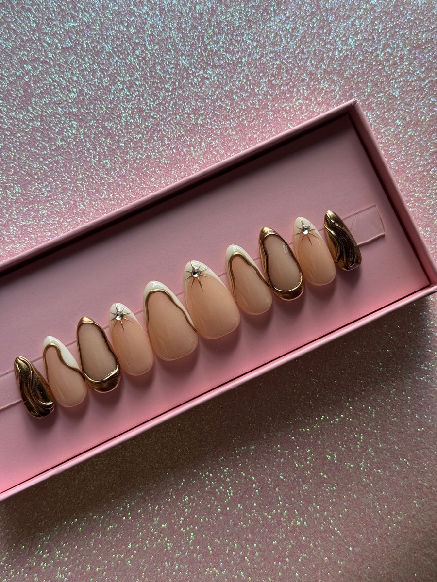 Set of almond-shaped press-on nails. Nails have a nude base, white tips, and gold trim. Some nails have a gold metallic finish and a small rhinestone star.