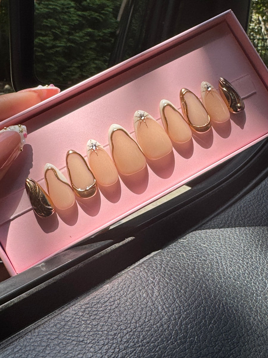 Short almond-shaped press-on nails with a nude base and white tips, outlined in gold. Some nails are embellished with star accents.