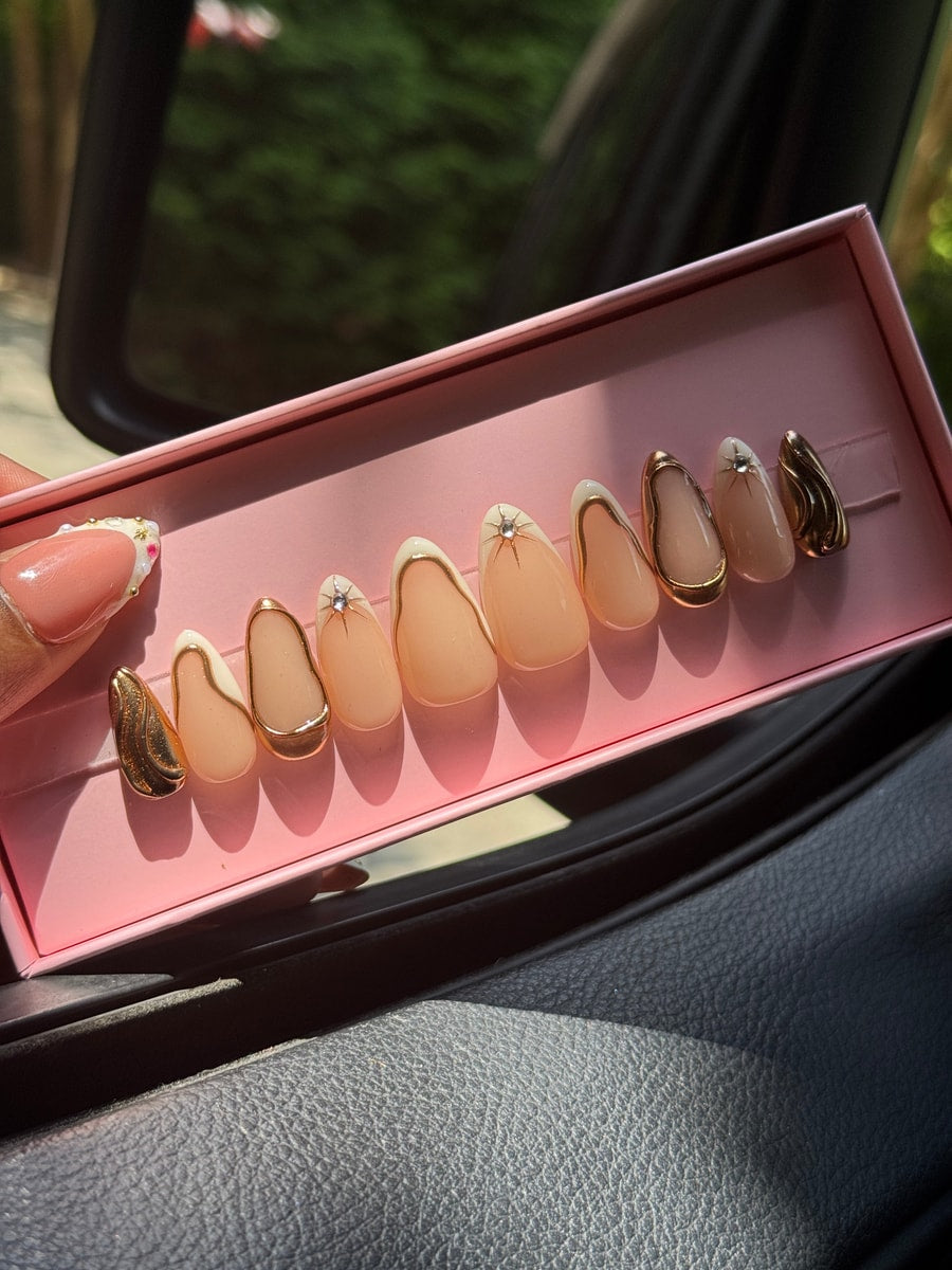 Almond-shaped super short press-on nails with nude, white, and gold accents, including gold outlines, geometric designs, and gem details. Some nails are fully gold.