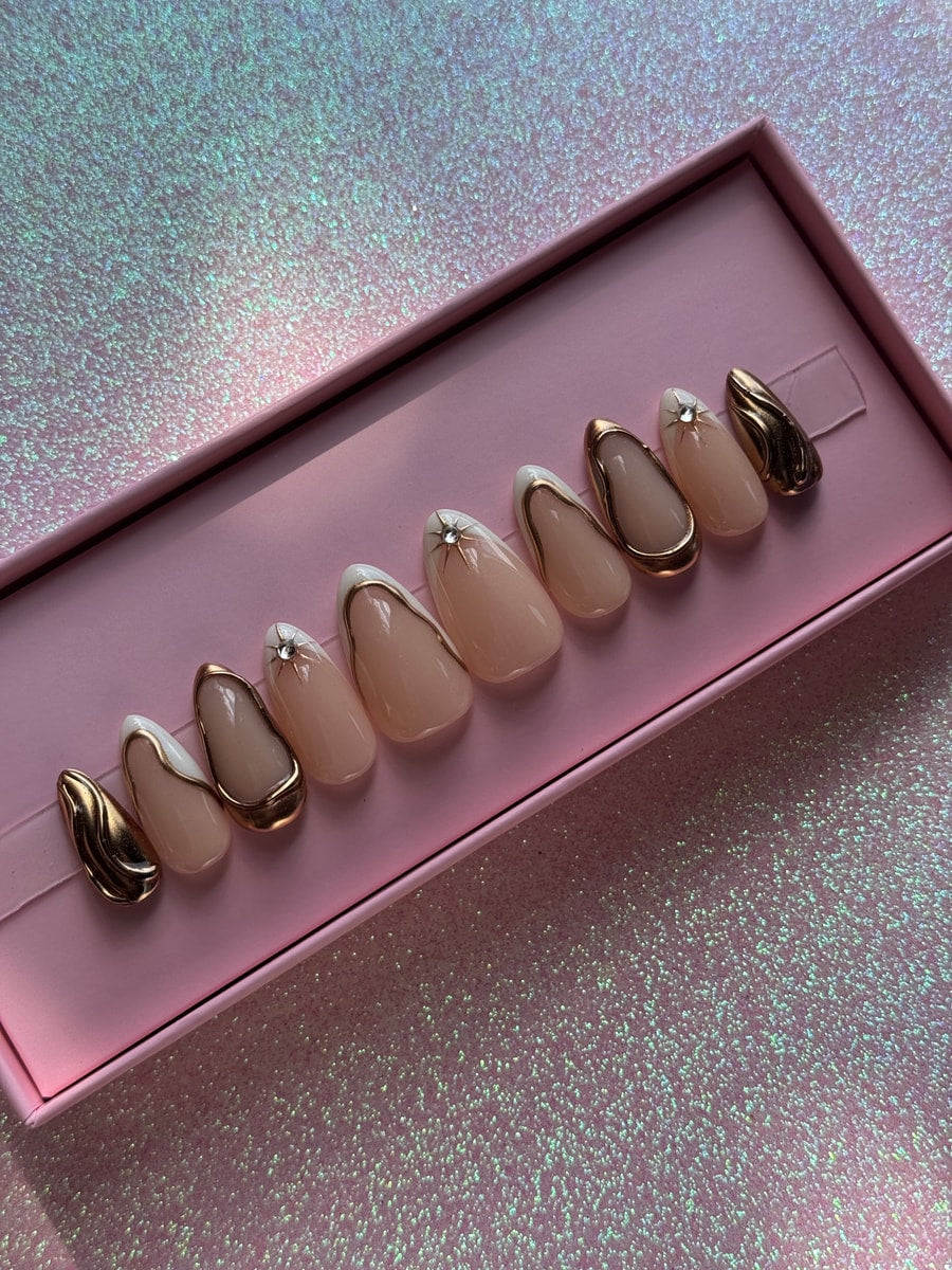 A set of super short almond-shaped press-on nails in a nude color with gold and white accents. Some nails have star embellishments while others have gold wave designs.