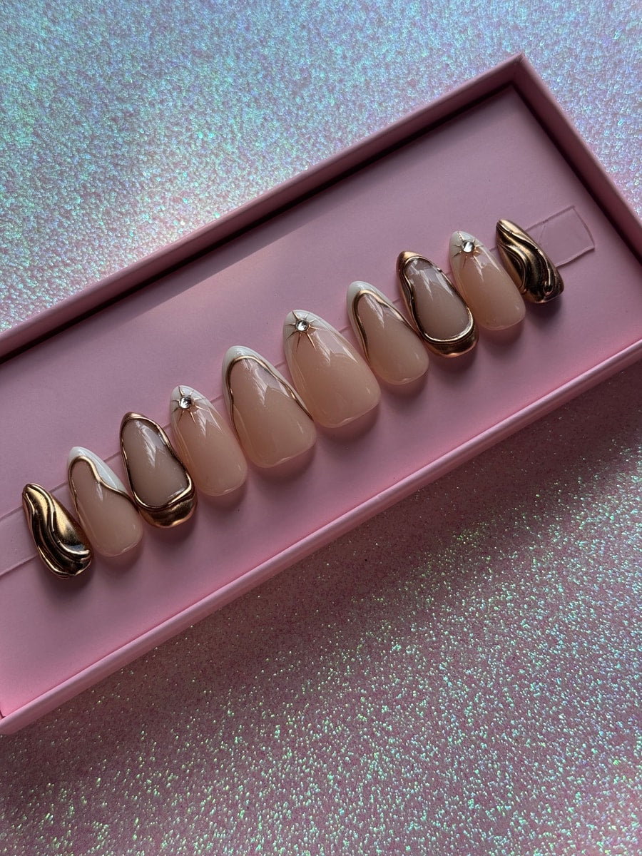 Super short almond shaped press-on nails with nude, white, and gold detailing. Some nails feature a rhinestone accent, and others are fully gold with a wavy texture.