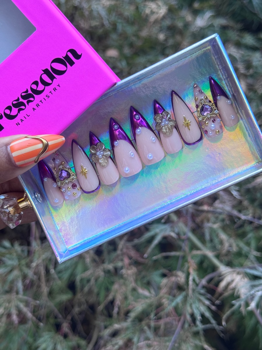 Stiletto-shaped press-on nails feature a light base color with a purple tip, pearl embellishments, and small gold floral charms.