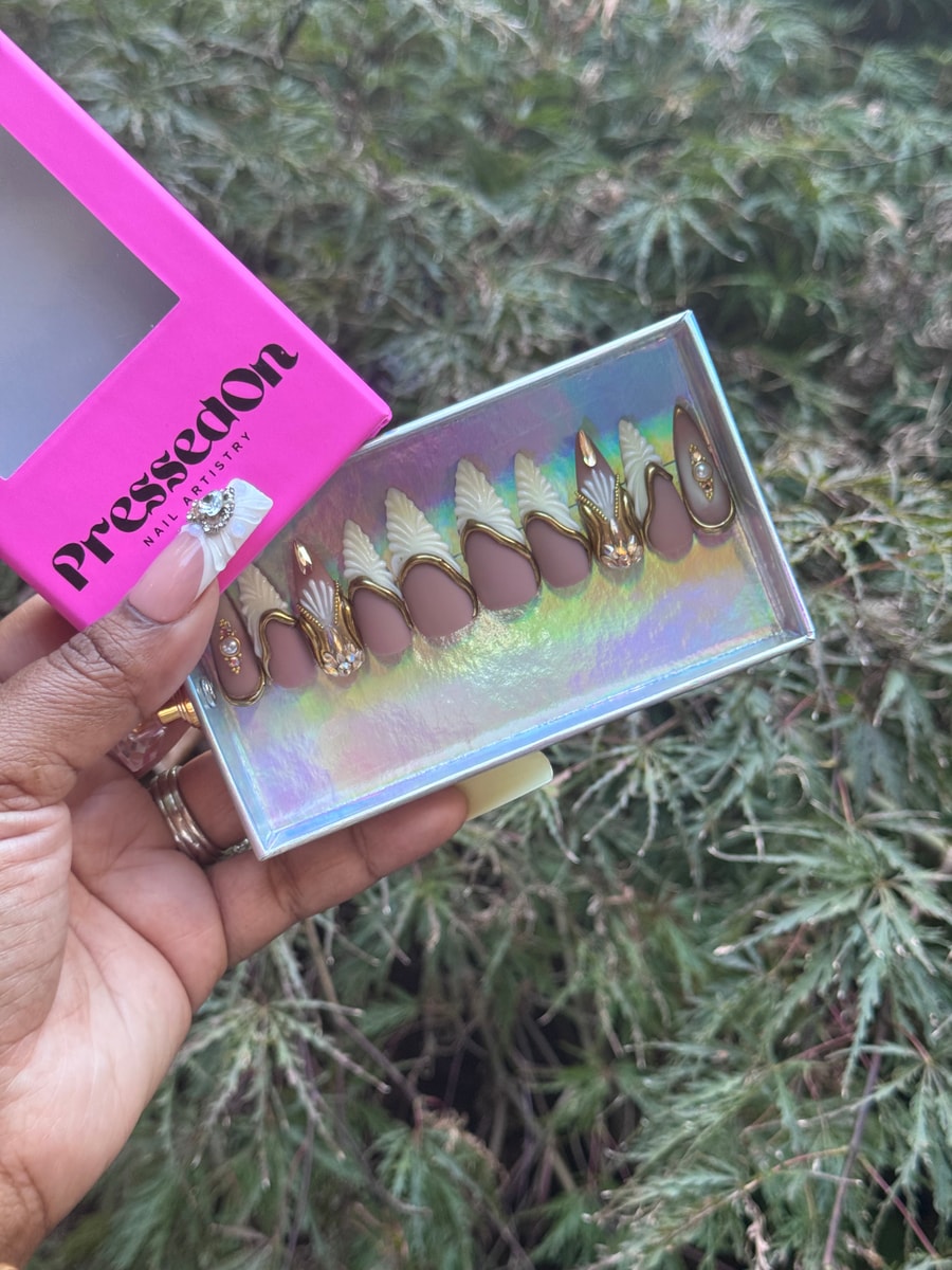 Medium-length press-on nails with a mauve base, gold detailing, white accents, rhinestones, and pearl embellishments.