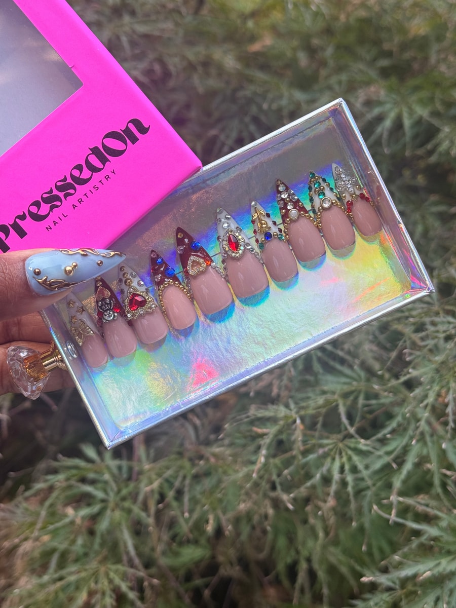 This set of short press-on nails features a nude base with rhinestone accents, gold details, and jewel-toned tips in red, green, and white. Some nails showcase heart or rose designs.
