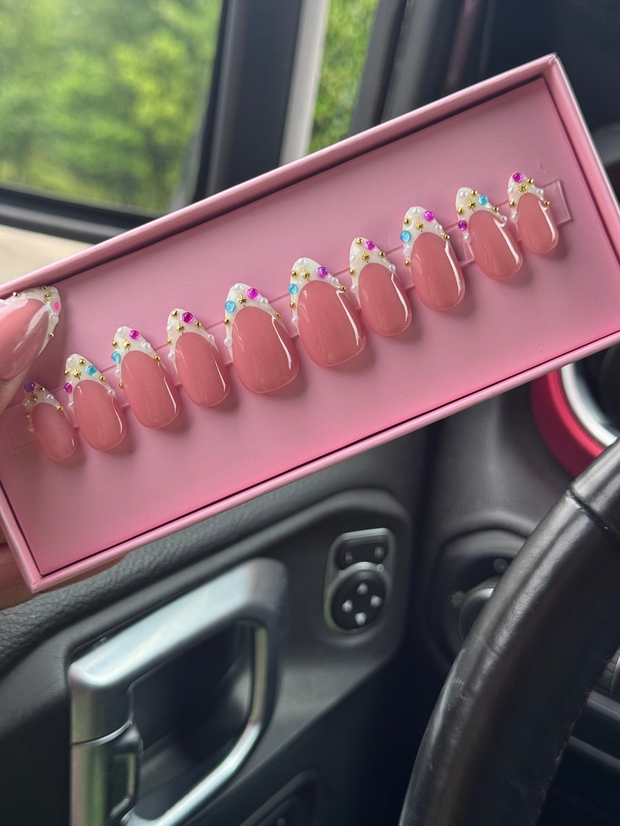 Light pink, super short press-on nails with white French tips. The tips are adorned with gold star embellishments, pearls, and small colorful gems.
