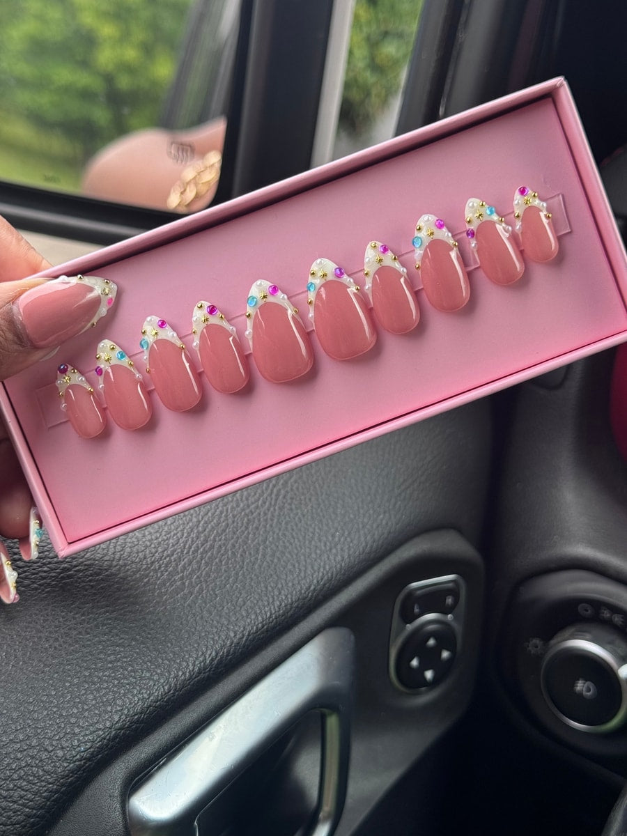 Light pink super short press-on nails with a white tip are decorated with gold stars, pearls, and colorful rhinestones.