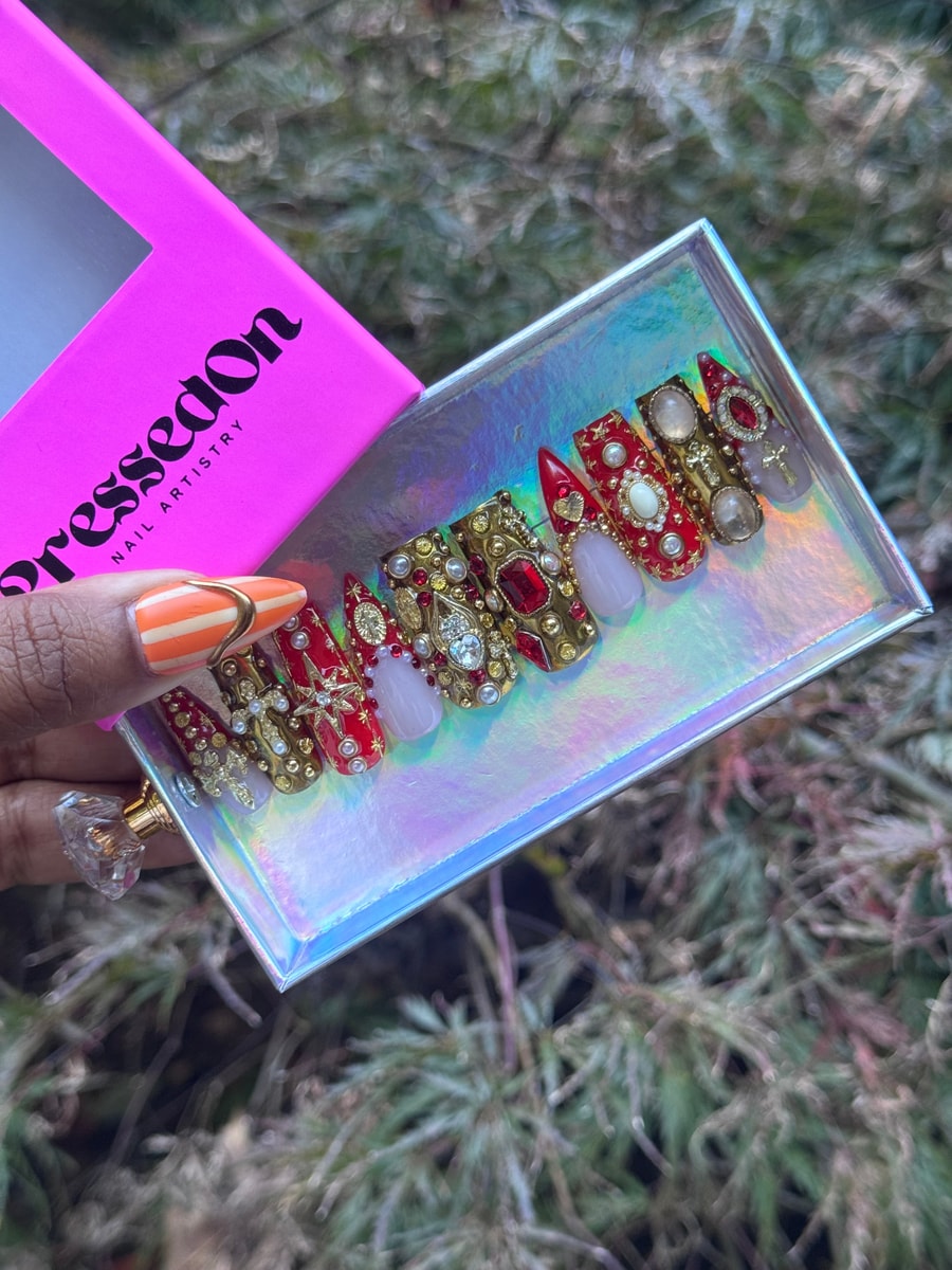 Ornate, medium-length press-on nails feature red and gold tones, enhanced with pearl and jewel accents and clear and ivory nails.